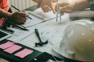 Blick auf Schreibtisch mit Bauplanungsskizzen, Werkzeug, Helm und Hände zweier Personen, die eine Planung besprechen